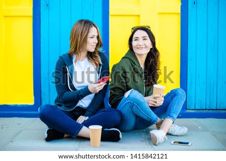 Two girls sitting on the ground with coffee to go and smartphones in their hands