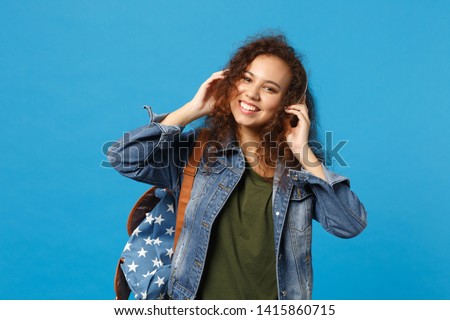 Young african american girl teen student in denim clothes, backpack headphones isolated on blue background studio portrait. Education in high school university college concept. Mock up copy space