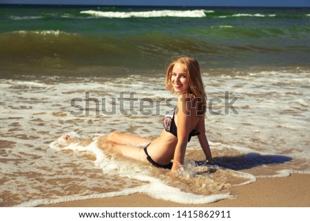 young blond woman in swimsuit at sea surf with foam sitting under sun rays, looking at camera