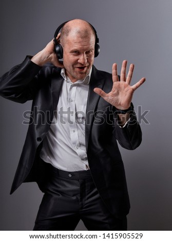 Happy emotional dancing and singing bald man listening the music in wireless headphone on dark grey background and showing five fingers on the palm. Closeup portrait
