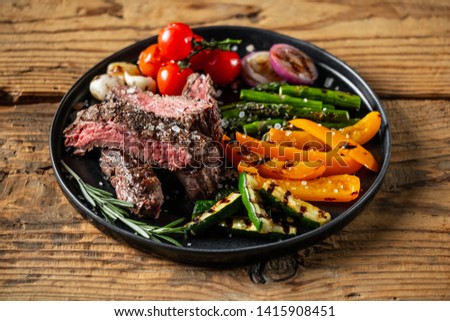 Grilled and sliced beef steak with grilled vegetables served on black plate on old rustic wood background.