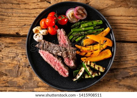 Grill menu concept - sliced beef steak with grilled vegetables served on black plate on old rustic wood background.