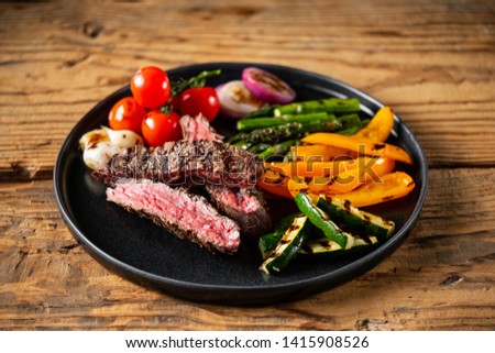 Sliced beef steak with grilled vegetables served on black plate on old rustic wood background.