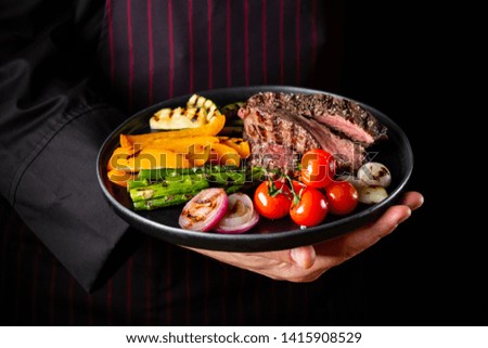 Grilled and sliced beef steak with grilled vegetables served on black plate on black background.