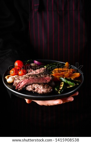Grilled and sliced beef steak with grilled vegetables served on black plate on black background.