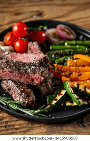 Grilled and sliced beef steak with grilled vegetables served on black plate on old rustic wood background.