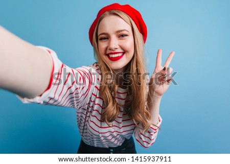 Joyful french girl taking selfie on blue background. Studio shot of laughing blonde lady showing peace sign.