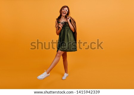 Full-length picture of girl having fun and taking photos on camera. Lady holds brown cardigan behind her back and walks on orange background