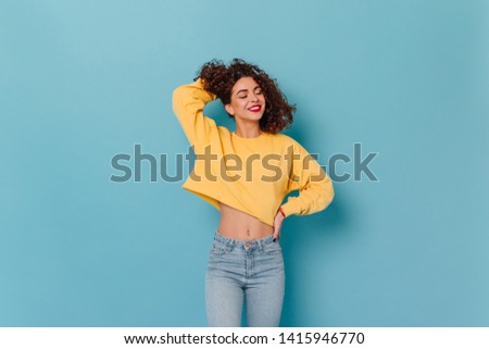 Girl touches her curly hair and smiles with her eyes closed. Photo of woman with red lipstick dressed in yellow sweater and denim pants on blue background