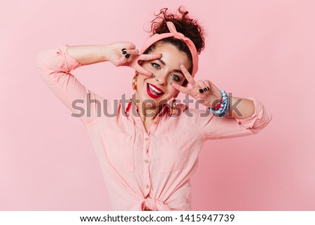 Blue-eyed girl with red lipstick shows signs of peace. Woman in headband and with bracelets on her arm is smiling on pink background