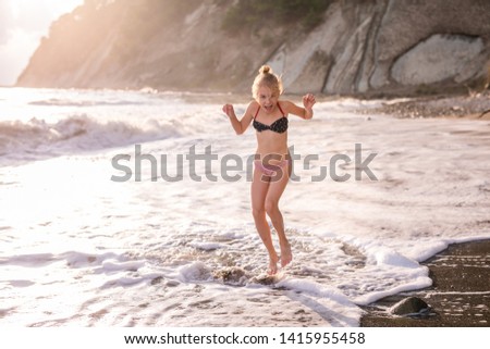 Adorable happy blonde girl in the white, red and blue swimming suit on beach vacation. Blue sea with white waves on the background.