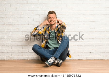 Blonde man sitting on the floor making phone gesture and pointing front