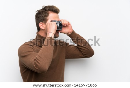 Blonde man over isolated white wall holding a camera
