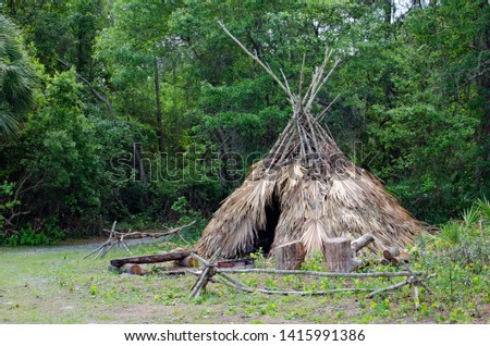 Camp setting of Seminole or Caribe Indian tiki hut tent made of palm fronds tied together with sticks and vines sitting behind a log bench and fire pit, and branch fences with green wooded background.