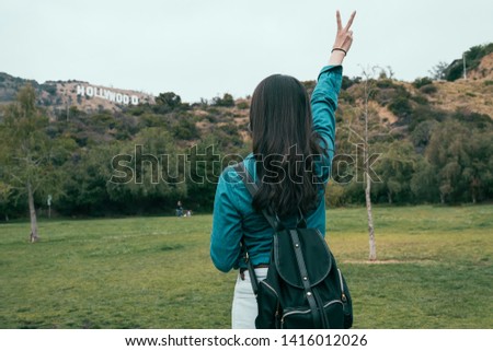 back view of young asian girl backpacker standing outdoor on summer lawn raise one hand to sky show victory win sign. female hiker achievement relax on hill taking photo with hollywood sign in park