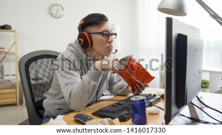 funny asian man with headphones plays video games on computer and eats snack from pack. japanese otaku gamer in glasses enjoy chips while watching anime cartoon movie on pc screen on summer break.