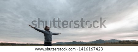 Wide view image of young man celebrating life standing with his arms spread widely under a glorious cloudy sky.