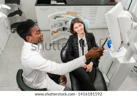Young African male dentist shows x-rays on the monitor. Woman in the dentist chair at dental clinic. Medicine, health, stomatology concept. dentist treating a patient.