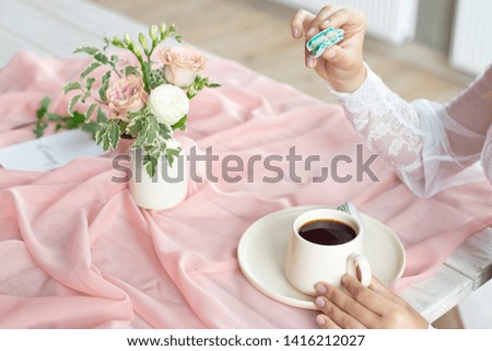 Young beautiful caucasian bride enjoying breakfast from french macaroon and coffee on a wooden table with a chiffon pink tablecloth and a vase of flowers.