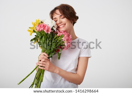 Cute young short haired girl in white blank t-shirt, holding a bouquet of colorful flowers, enjoying the smell, broadly smiling with closed eyes, standing over white background.