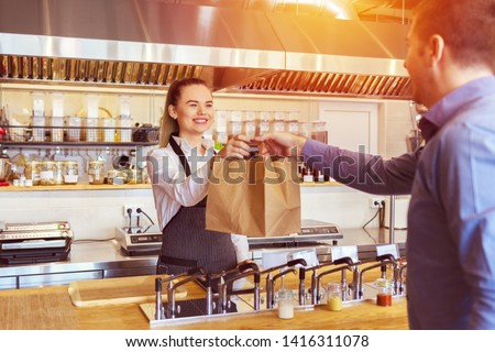 Cheerful waitress wearing apron serving customer at counter in restaurant - Small business and service concept with young woman owner offering recycled paper bag with take away food to online client Royalty-Free Stock Photo #1416311078