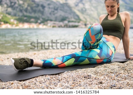 Young sportive woman doing yoga on mat, exercising on the beach. Girl meditate by the coast, enjoying relaxation. Doing leg stretching