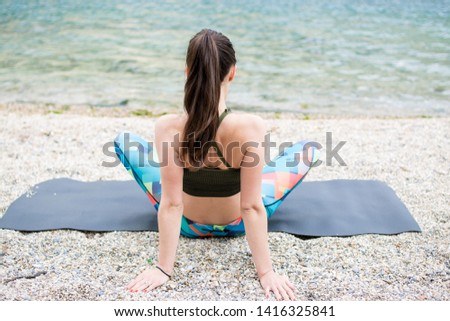Young beautiful sportive woman sitting on yoga mat, exercising on the beach. Girl meditate by the coast, enjoying the relaxation. Doing yoga by the sea