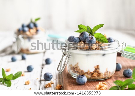Healthy breakfast of blueberry parfaits made with fresh fruit, Greek yogurt, granola and mint leaves over a rustic white table. Selective focus on glass jar in front with blurred background.
