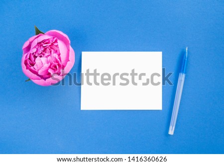 Flat lay of beautiful pink peony flower on blue background with pen, and white isolated paper.