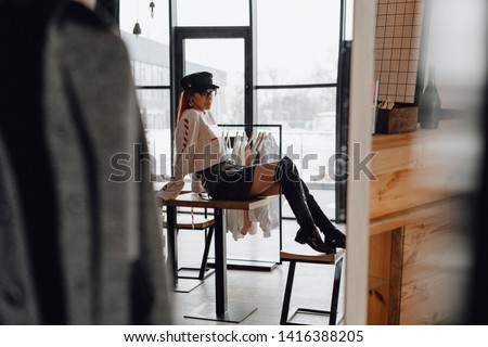 Beautiful Woman in Fashion Store Owner or Designer. Stylish Redhead Wearing Trendy Sweatshirt and High Boots Sitting at Table. Rack with Apparel at Modern Boutique. Natural Light from Window