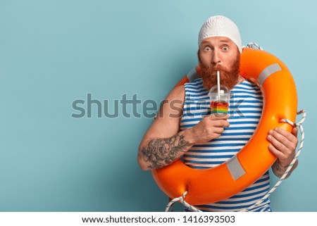 Embarrassed holiday maker drinks cold summer cocktail, spends free time at beach, wears swimcap sailor t shirt, swims with lifebuoy, has surprised expression, models over blue wall with free space Royalty-Free Stock Photo #1416393503