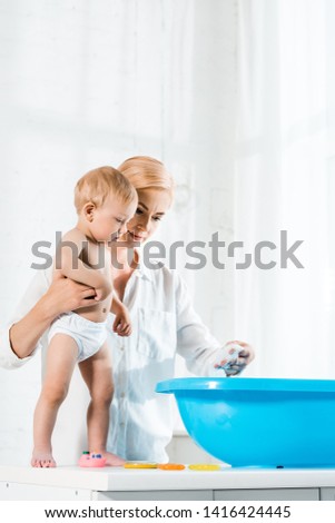low angle view of attractive woman holding toddler son while standing near blue baby bathtub 