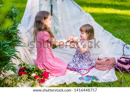 Two sisters in multi-colored dresses sit in white boho tent outdoor and play in finger game.