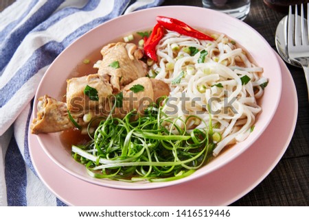 Famous noodle and chicken soup served with spoon and fork on a rose plate, green onions stripes, red chili pepper slices, on a dark wooden background, close-up 