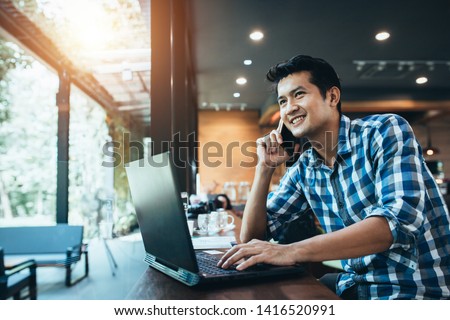Asian man freelance work on computer touch pad while talking on smart phone With a happy smile, young business man use laptop sitting at wooden table of modern coffee shop, freelance man.