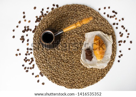 Creative flat lay  with straw woven round as background,  cezve with fresh hot coffee, croissant and pieces of chocolate at parchment, copy space and chaotic coffee beans at the white background