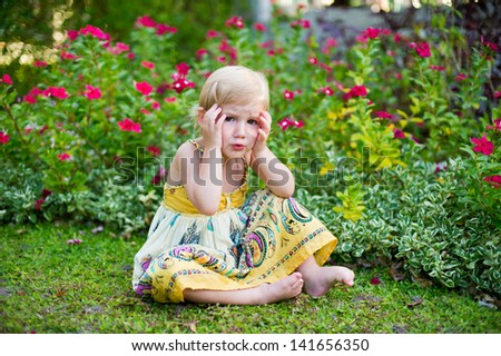 Adorable scared little girl in dress sitting in tropical blooming garden and holding her head