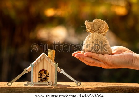 Man hand holding money bag with submit to a wooden home and construction tools with repairing on sunlight in the public park, Saving money and loan for renovation to real estate and house concept.