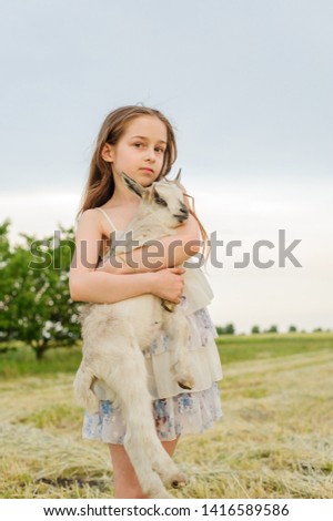 Little girl plays and huhs goatling in country, spring or summer nature outdoor. Cute kid with baby animal, countryside, forest, trod, glade background. Friendship of child and yeanling, image toned.