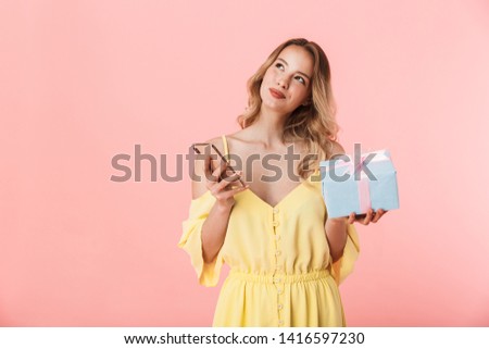 Image of a beautiful excited emotional happy young blonde woman posing isolated over pink wall background holding present gift box using mobile phone.