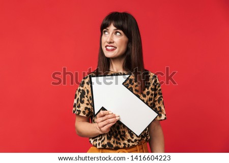 Photo of beautiful brunette woman 30s dressed in stylish outfit smiling and holding blank arrow pointer isolated over red background
