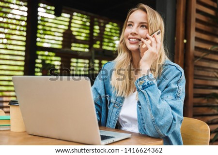 Image of amazing excited happy young woman posing outdoors in cafe using laptop computer and talking by mobile phone.
