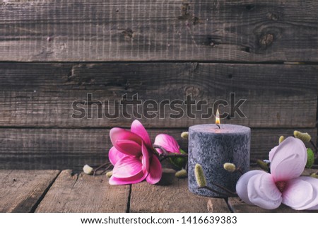 Still life wellness concept. Black burning candle  and pink magnolia flowers on  wooden rustic  background.  Selective focus. Place for text. Toned image.
