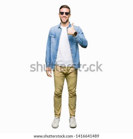 Handsome man wearing fashion sunglasses doing happy thumbs up gesture with hand. Approving expression looking at the camera with showing success.