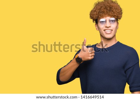 Young handsome man with afro wearing glasses doing happy thumbs up gesture with hand. Approving expression looking at the camera with showing success.