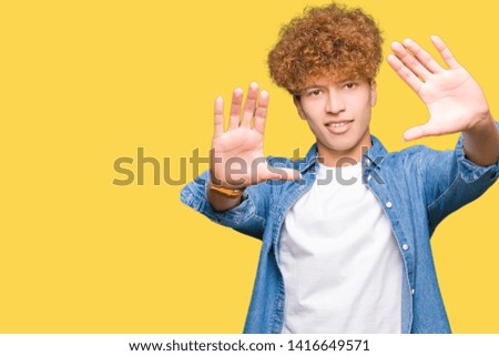 Young handsome man with afro hair wearing denim jacket Smiling doing frame using hands palms and fingers, camera perspective