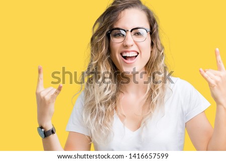Beautiful young blonde woman wearing glasses over isolated background shouting with crazy expression doing rock symbol with hands up. Music star. Heavy concept.