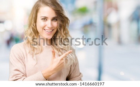 Beautiful young blonde woman wearing sweatershirt over isolated background cheerful with a smile of face pointing with hand and finger up to the side with happy and natural expression on face