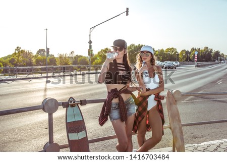 Two girls girlfriends students drink water in summer city, skate boards, swimsuit shorts. Resting long hair tanned figure. Background road cars. Fashion style, modern lifestyle. Free space for text