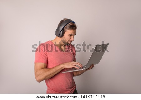 Man in wireless headphone playing games online on portable notebook gadget while standing isolated in studio against wall with copy space.  